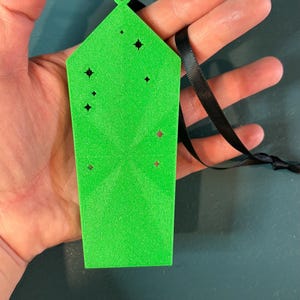 Puede incluir: Expositor de pendientes verde brillante, rectangular, con recortes en forma de estrella y una cinta negra. El expositor se sostiene en una mano, mostrando su color y dise&ntilde;o. Un accesorio &uacute;nico.