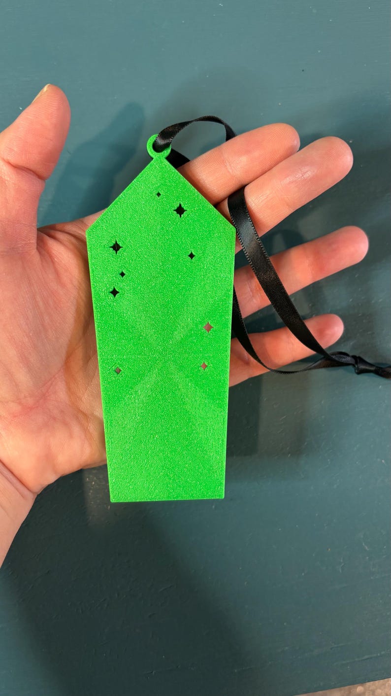 Puede incluir: Expositor de pendientes verde brillante, rectangular, con recortes en forma de estrella y una cinta negra. El expositor se sostiene en una mano, mostrando su color y dise&ntilde;o. Un accesorio &uacute;nico.