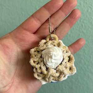 May include: A miniature dessert charm featuring a white swirl of frosting atop a beige, textured pastry base. The charm is attached to a silver chain and a wooden ring. The charm is held in a hand against a light green background.