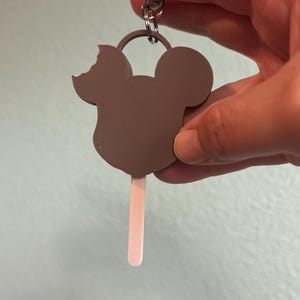 Puede incluir: Un llavero de paleta con forma de Mickey Mouse marr&oacute;n con una mordida en la oreja. El palito de la paleta es de color rosa claro. El llavero se sujeta con un anillo plateado.