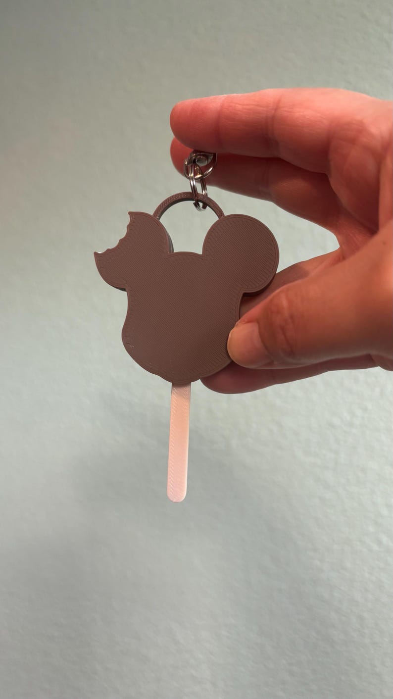 Puede incluir: Un llavero de paleta con forma de Mickey Mouse marr&oacute;n con una mordida en la oreja. El palito de la paleta es de color rosa claro. El llavero se sujeta con un anillo plateado.