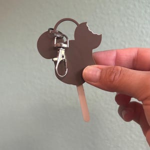 Puede incluir: Un llavero marr&oacute;n con forma de Mickey Mouse con un mordisco en el helado. El llavero est&aacute; sujeto a un cierre y un anillo plateados. El palito del helado es marr&oacute;n claro. El fondo es verde claro liso.