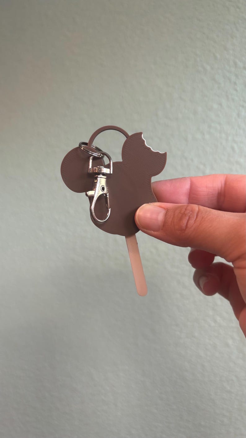 Puede incluir: Un llavero marr&oacute;n con forma de Mickey Mouse con un mordisco en el helado. El llavero est&aacute; sujeto a un cierre y un anillo plateados. El palito del helado es marr&oacute;n claro. El fondo es verde claro liso.