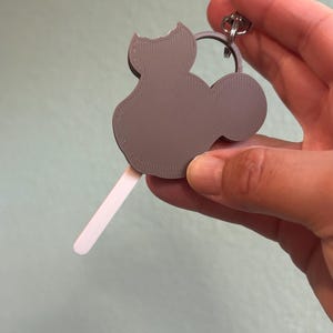 Puede incluir: Un llavero gris con forma de gato con una mordida en la oreja, sujeto a un llavero plateado. El llavero tiene un palito de helado adjunto en la parte inferior. El llavero se sostiene en una mano.