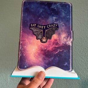 May include: A tablet case with a galaxy design and the words "BAT SHIT CRAZY" above a bat illustration. The case is held in a white and blue stand. The background is a solid, muted green.