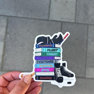 May include: A sticker featuring a stack of books with titles like "Goldeneyes" and "Fleet," topped with hockey gloves and a hockey stick. A pair of black ice skates is on the right. The sticker is held by a hand.