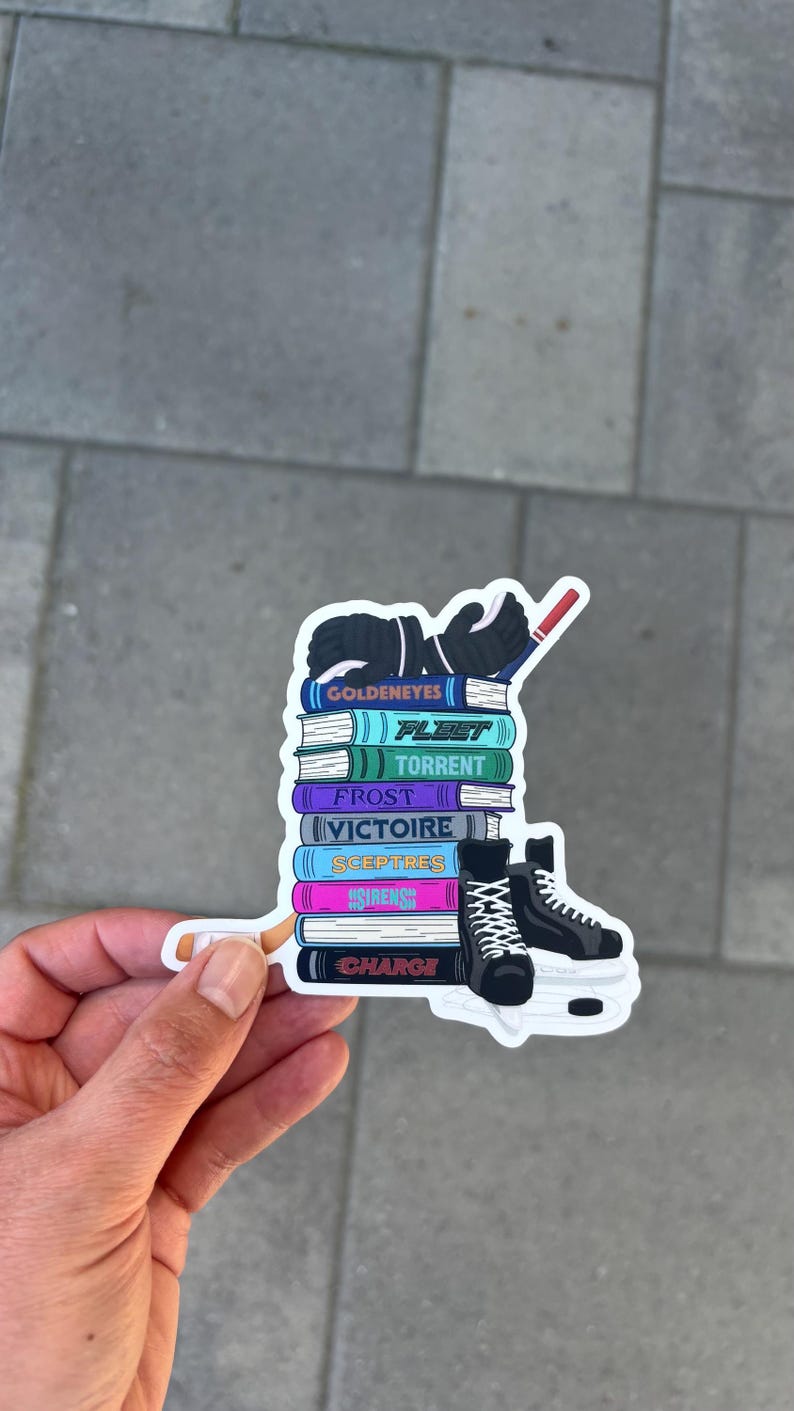 May include: A sticker featuring a stack of books with titles like "Goldeneyes" and "Fleet," topped with hockey gloves and a hockey stick. A pair of black ice skates is on the right. The sticker is held by a hand.