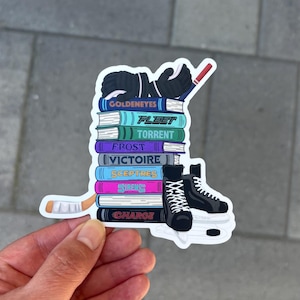 May include: A sticker featuring a stack of books with hockey-themed elements. The books have titles like "Goldeneyes" and "Fleet." Hockey gloves, a hockey stick, and ice skates are also depicted. The sticker is held by a hand.