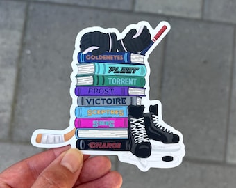 Puede incluir: Una pegatina con una pila de libros con elementos temáticos de hockey. Los libros tienen títulos como "Goldeneyes" y "Fleet". También se representan guantes de hockey, un palo de hockey y patines. La pegatina está sujeta por una mano.