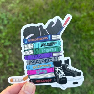 May include: A sticker featuring a stack of books with hockey-themed titles like "Goldeneyes" and "Fleet." The design includes hockey gloves, skates, and a hockey stick. The sticker is held by a hand.