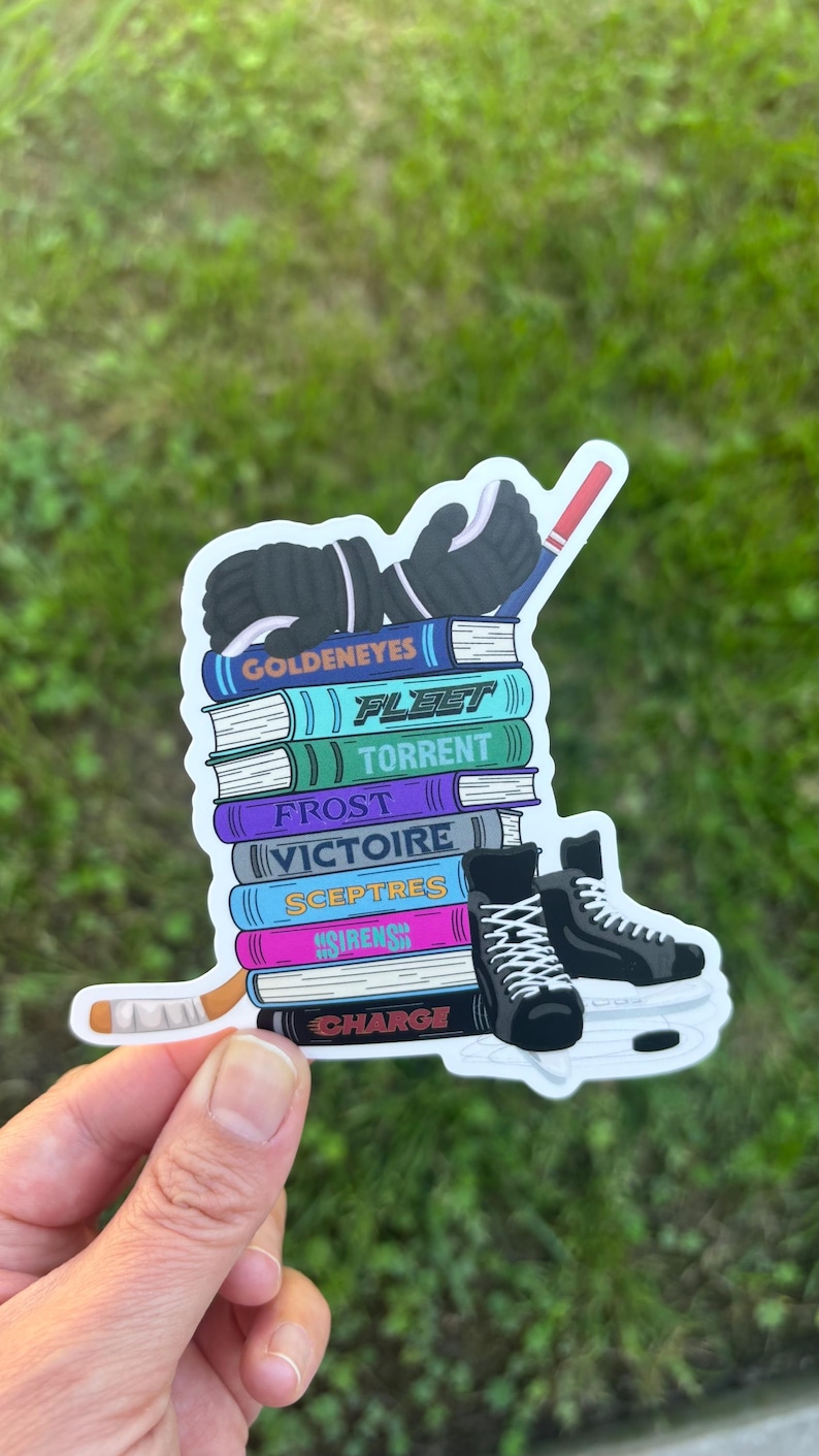 May include: A sticker featuring a stack of books with hockey-themed titles like "Goldeneyes" and "Fleet." The design includes hockey gloves, skates, and a hockey stick. The sticker is held by a hand.