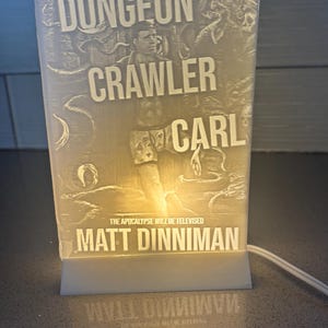 May include: A rectangular, illuminated lamp featuring the text "DUNGEON CRAWLER CARL" and the author's name, Matt Dinniman. The lamp is white and emits a soft glow, with the text and design etched into the surface.