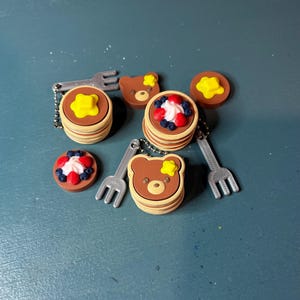 May include: A collection of miniature pancake charms with a bear face design, topped with butter, whipped cream, and berries. Each charm is attached to a small silver fork. The pancakes are stacked and have a light brown color.