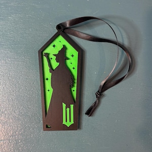 Puede incluir: Adorno negro y verde con tem&aacute;tica de brujas con una cinta negra. El adorno presenta una silueta de bruja y la letra "W". El fondo es verde con detalles de estrellas.