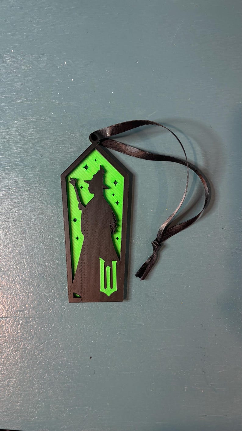 Puede incluir: Adorno negro y verde con tem&aacute;tica de brujas con una cinta negra. El adorno presenta una silueta de bruja y la letra "W". El fondo es verde con detalles de estrellas.