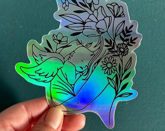 May include: A holographic sticker featuring a black line drawing of a woman holding a child, surrounded by flowers. The sticker has a rainbow iridescent finish.
