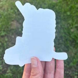 May include: A white sticker in the shape of a semi-truck. The sticker is held in a hand against a background of green grass. The truck shape has a detailed cab and trailer design, with a distinct outline.