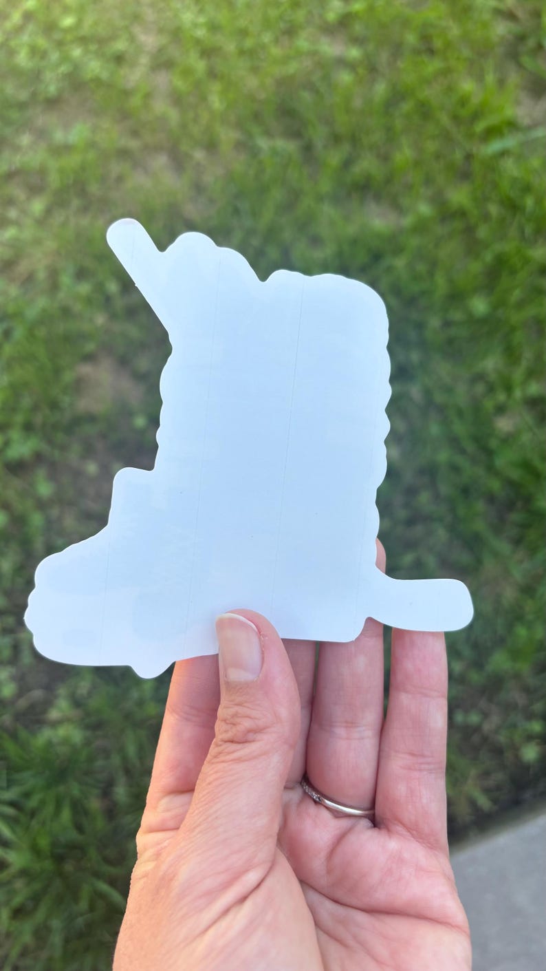 May include: A white sticker in the shape of a semi-truck. The sticker is held in a hand against a background of green grass. The truck shape has a detailed cab and trailer design, with a distinct outline.