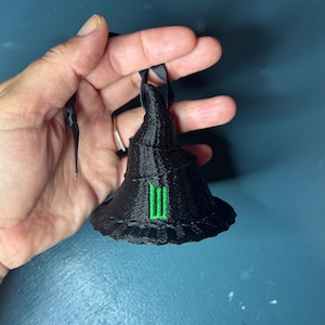May include: A black witch hat ornament with a green emblem. The hat is made of a textured material and has a black ribbon for hanging. A festive decoration for Halloween or other occasions.