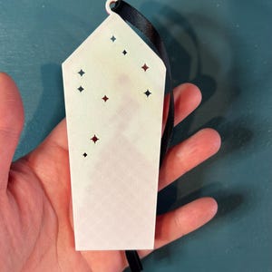 Puede incluir: Un adorno rectangular blanco con recortes en forma de estrella y una cinta negra para colgar. El adorno tiene una superficie ligeramente texturizada y se sostiene sobre un fondo turquesa. Los recortes est&aacute;n acentuados con formas de estrella rojas y negras.