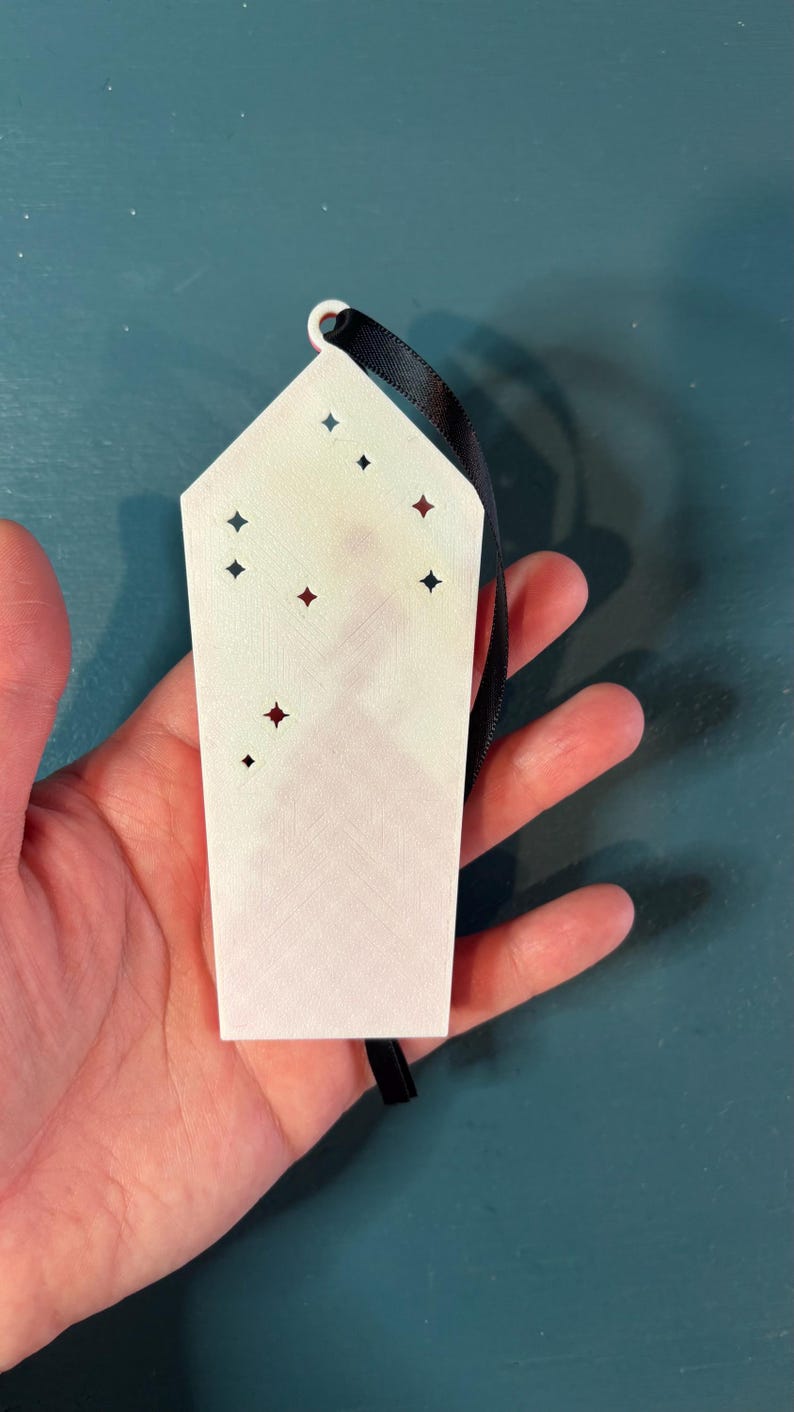 Puede incluir: Un adorno rectangular blanco con recortes en forma de estrella y una cinta negra para colgar. El adorno tiene una superficie ligeramente texturizada y se sostiene sobre un fondo turquesa. Los recortes est&aacute;n acentuados con formas de estrella rojas y negras.