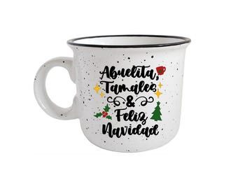 Puede incluir: Taza de cerámica blanca con motas negras y borde negro. La taza presenta el texto "Abuelita, Tamales & Feliz Navidad" con ilustraciones festivas. Un asa está unida al lateral de la taza.