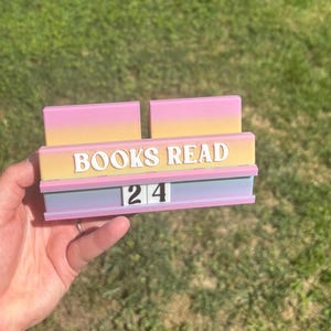 May include: A pastel-colored book tracker with the words "BOOKS READ" in white lettering. The number "24" is displayed below. The tracker is held against a background of green grass.