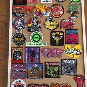 May include: A collection of embroidered patches featuring various band logos and album art, including Nirvana, AC/DC, and The Doors. The patches are arranged on a corkboard with a white frame, showcasing a variety of colors and designs.