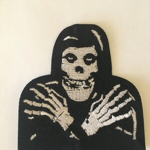 May include: Black and white embroidered patch of a skeleton with its arms crossed in front of its chest. The skeleton is wearing a black hooded robe.