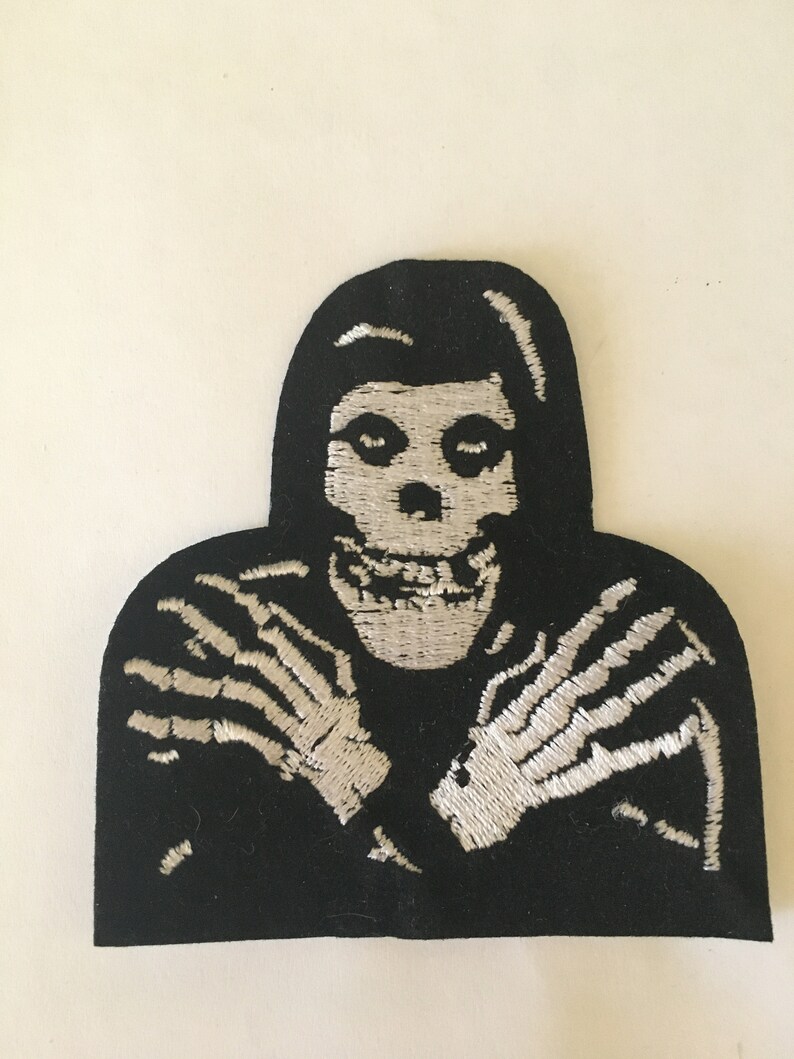 May include: Black and white embroidered patch of a skeleton with its arms crossed in front of its chest. The skeleton is wearing a black hooded robe.