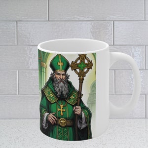 May include: White ceramic mug with a green, gold, and white illustration of Saint Patrick holding a staff with a shamrock on top. The background is a green and white illustration of a church.