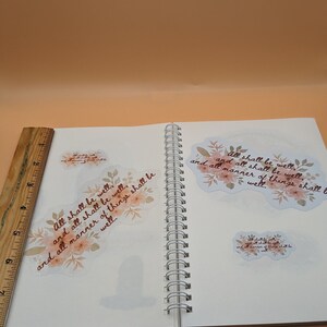 May include: A white notebook with a spiral binding. Two floral stickers with the text "All shall be well, and all shall be well, and all manner of things shall be well." are on the pages.
