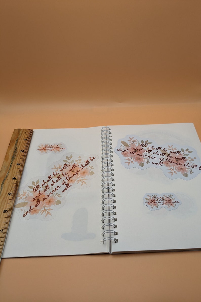 May include: A white notebook with a spiral binding. Two floral stickers with the text "All shall be well, and all shall be well, and all manner of things shall be well." are on the pages.