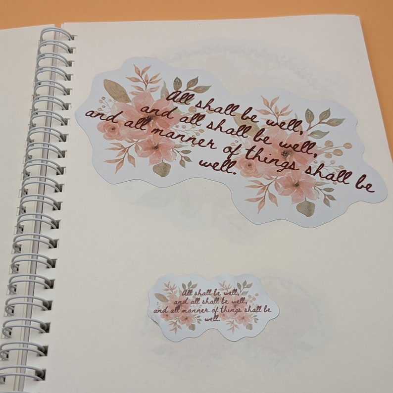 May include: A white page with two watercolor floral stickers. The stickers feature the text "All shall be well, and all shall be well, and all manner of things shall be well."