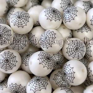 Peut inclure: Un ensemble de perles blanches et rondes ornées d'une toile d'araignée noire. Les perles ont une finition brillante et un trou pour l'enfilage. Le texte "Gladiolus Beading Supplies" est visible.
