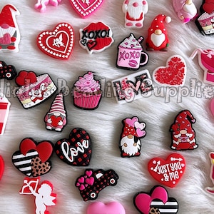 May include: A collection of Valentine's Day-themed charms in various shapes and colors. The charms include hearts, gnomes, cupcakes, and coffee cups. Some charms have text like "Love," "XOXO," and "I love you." The charms are on a white, fluffy surface.