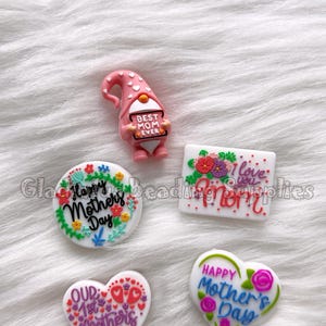 May include: Five colorful resin Mother's Day themed embellishments.  A pink gnome holding a sign that says "Best Mom Ever". A white heart with the text "Our 1st Mother's Day". A white circle with the text "Happy Mother's Day". A white rectangle with the text "Mom I love you". A white heart with the text "Happy Mother's Day".