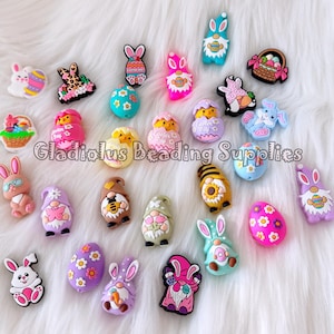 May include: A selection of colourful Easter-themed charms, including bunnies, chicks, gnomes, and decorated eggs. The charms are in various colours and designs, some with floral patterns and others with glitter accents. The text "Gladiolus Beading Supplies" is visible.