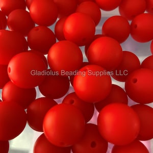 May include: A close-up image of a large quantity of bright red silicone beads. The beads are smooth and round, with a small hole in the center of each bead. The beads are arranged in a random pattern, creating a visually appealing display.