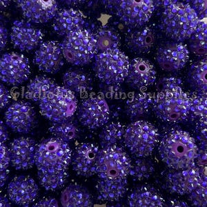 May include: A close-up of a pile of purple beads covered in small, sparkly blue rhinestones. The beads are round and have a hole in the centre for stringing.
