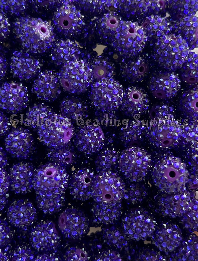 May include: A close-up of a pile of purple beads covered in small, sparkly blue rhinestones. The beads are round and have a hole in the centre for stringing.
