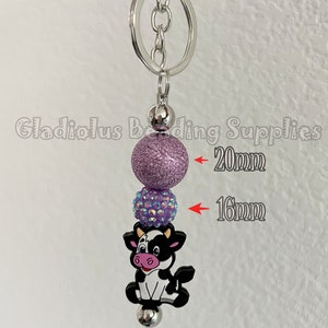 May include: A purple and iridescent charm keyring with a cow charm. The purple bead is 20 mm in diameter and the iridescent bead is 16 mm in diameter.