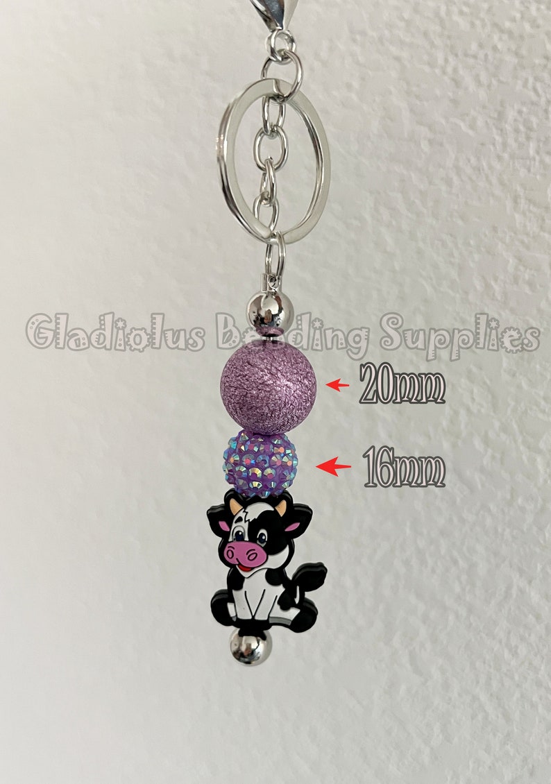 May include: A purple and iridescent charm keyring with a cow charm. The purple bead is 20 mm in diameter and the iridescent bead is 16 mm in diameter.