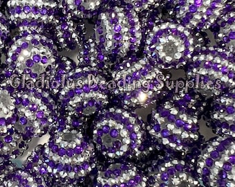 May include: A close-up of a group of purple and silver rhinestone beads. The beads are round and have a sparkly finish.