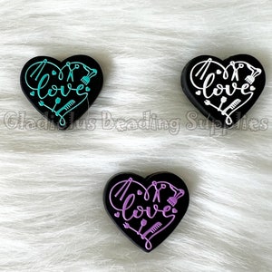 May include: Three black heart-shaped charms with the word "love" written in a stylized font. Each charm features different colored hair styling tools, including scissors, combs, and hair dryers. The charms are on a white fuzzy background.