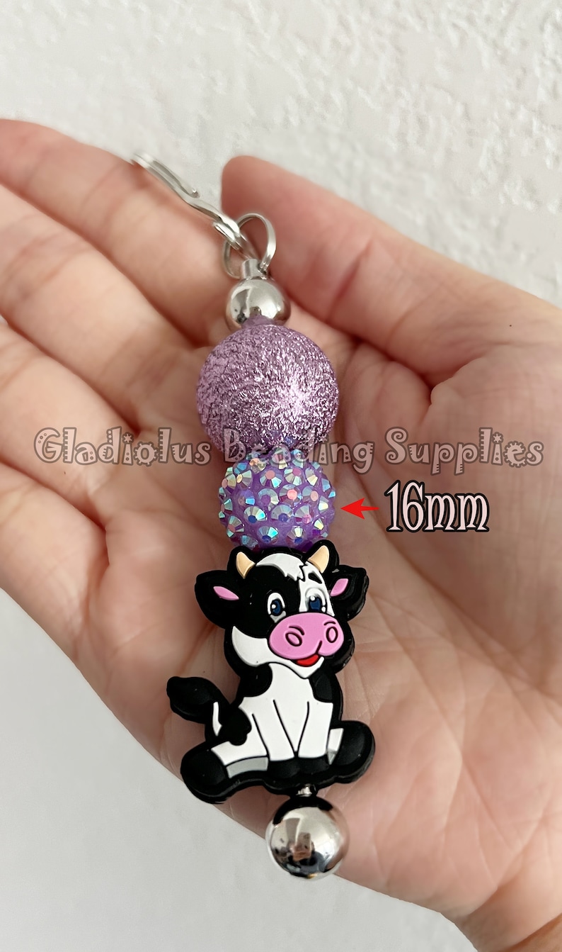 May include: A purple glitter bead, a purple iridescent bead, and a black and white cow charm with pink accents on a silver chain. The cow charm is 16mm in size.