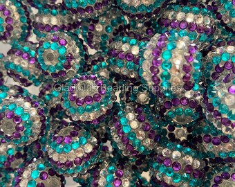 May include: A close-up of a pile of round, iridescent beads. The beads are covered in small, clear rhinestones and have a teal and purple color scheme.  The beads are perfect for jewelry making or crafting.