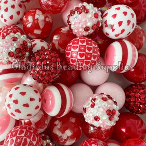 May include: A collection of red and pink Valentine's Day themed beads. The beads are made of plastic and have various designs, including hearts, stripes, and polka dots. Some beads are covered in rhinestones.