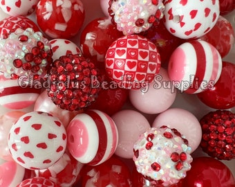 May include: A collection of red and pink Valentine's Day themed beads. The beads are made of plastic and have various designs, including hearts, stripes, and polka dots. Some beads are covered in rhinestones.