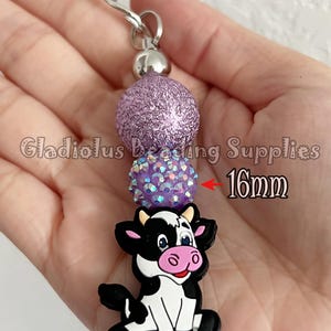 Puede incluir: Un llavero artesanal con un dije de vaca en blanco y negro, cuentas de purpurina moradas y un cierre plateado. Las cuentas miden 1,6 cm. El texto "Gladiolus Beading Supplies" es visible.